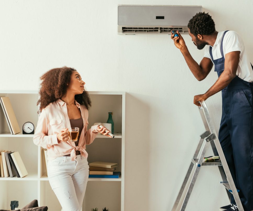 african american repairman fixing air conditioner near pretty african american woman holding remote controller
