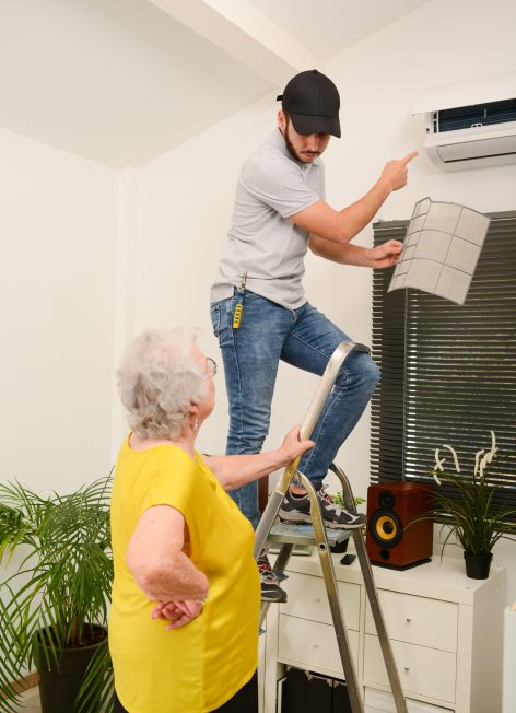 handsome young man electrician cleaning air filter on an indoor unit of air conditioning system in client house with senior woman
